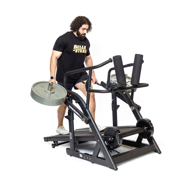 A man in a black shirt with "Bells of Steel" branding uses The Dreadmill equipped with a barbell rack for magnetic resistance training. With curly hair and beard, he enjoys an intense cardio workout while building strength.