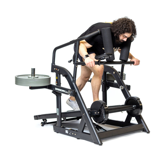 A person with curly hair uses Bells of Steel's The Dreadmill, showcasing the magnetic resistance. Bent over and gripping the handles, weights flank the sides. Wearing a black shirt and light-colored shoes, they add flair to their workout session.