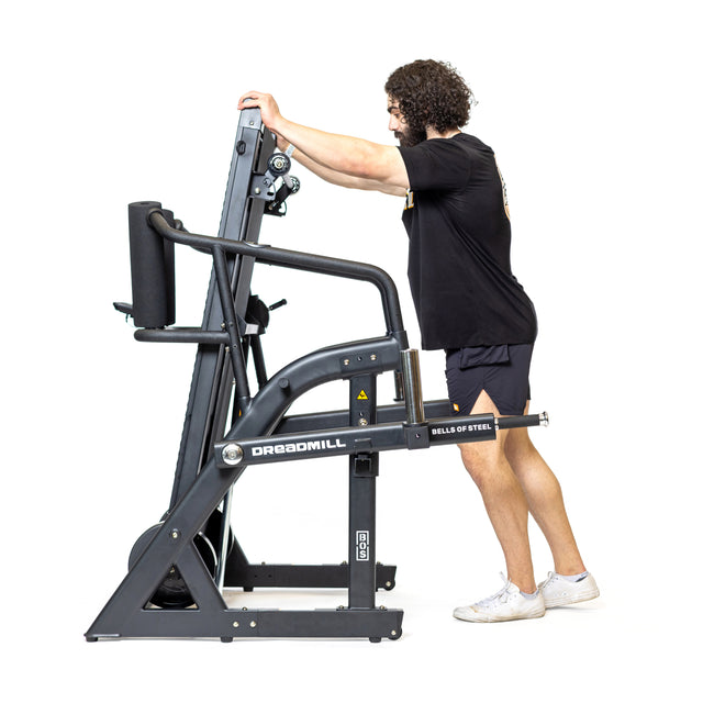 A person using The Dreadmill by Bells of Steel, dressed in a black shirt and shorts with white sneakers. They're gripping the handles, leaning forward in an energetic workout pose against a stark white background.