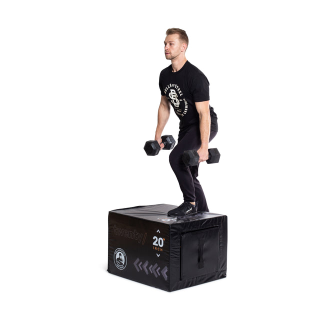 A man in black athletic wear performs a step-up with dumbbells on the Bells of Steel 6-In-1 Soft Foam Plyo Box/Squat Box, demonstrating its space-saving design.