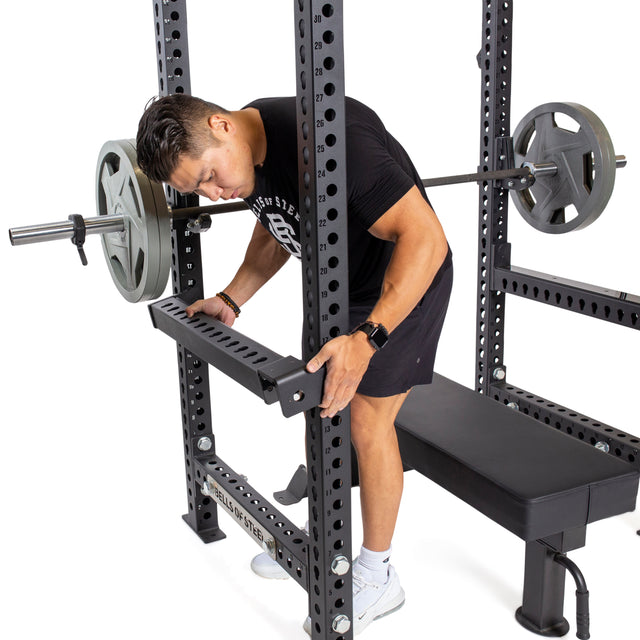 A person wearing a black shirt and shorts adjusts the Bells of Steel Flip-Down Safeties on a power rack with a loaded barbell beside a flat bench, ensuring optimal workout safety.
