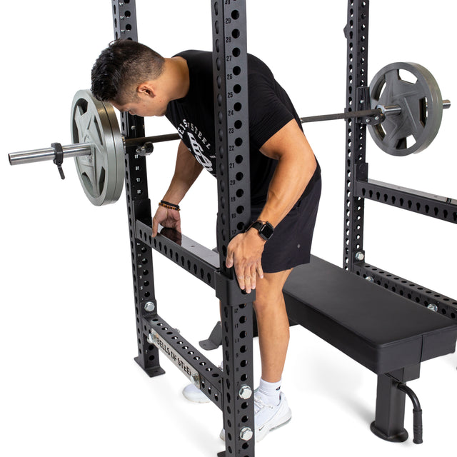 Wearing black athletic clothes, a person adjusts the Bells of Steel Flip-Down Safeties on a power rack to ensure training safety while setting up the bench and loaded barbell.