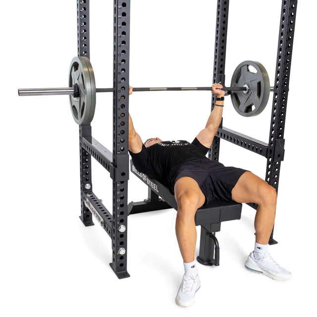 Wearing athletic clothing, a person bench presses with a barbell on a bench inside a power rack, using Bells of Steel Flip-Down Safeties for added safety during training.