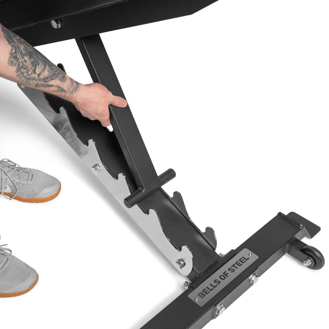 A person adjusts the incline of the Bells of Steel Flat / Incline / Decline Weight Bench - Commercial. Only their tattooed arm, hand, and lower legs in gray sneakers are visible, emphasizing this versatile workout bench.