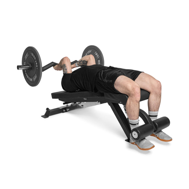 A person lies on the Bells of Steel Flat / Incline / Decline Weight Bench - Commercial with leg support, gripping a barbell and preparing to lift. They wear black workout clothes and gray sneakers.