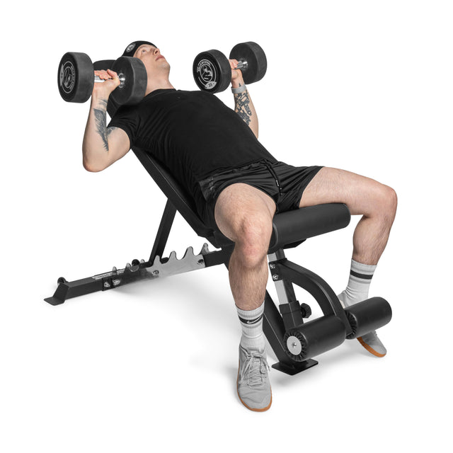 Wearing black workout clothes and a beanie, a person uses the Bells of Steel Flat / Incline / Decline Weight Bench - Commercial to perform an incline dumbbell press, lifting two dumbbells above their chest for targeted muscle engagement.