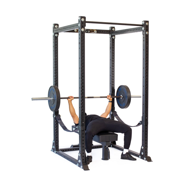 Using Bells of Steel's Hydra Flat Foot Components, a person bench presses with a barbell on a flat bench inside the sleek black rack that matches their black athletic attire perfectly.