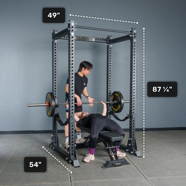 Two people use a Bells of Steel Manticore Flat Foot Power Rack - Prebuilt (3" x 3", 1" holes) for bench pressing in a home gym. The rack’s dimensions are marked: 49" wide, 54" deep, and 87¼" tall. One person is lying on a bench pressing a barbell while the other stands nearby spotting.