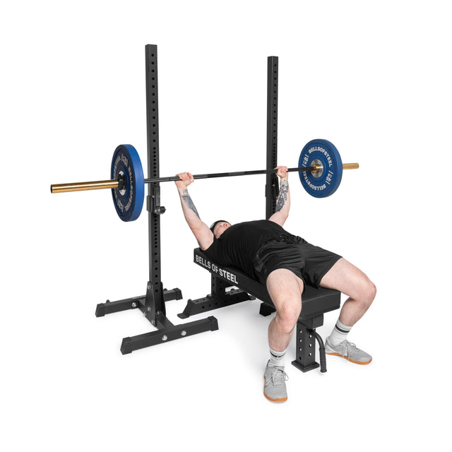 A person in black shorts, a black shirt, and gray sneakers lies on the Bells of Steel Powerlifting Flat Bench with durable vinyl, lifting a barbell with blue plates on a commercial-grade bench press rack.