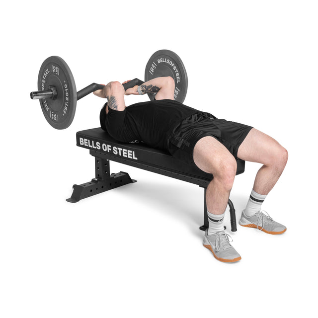 Wearing athletic gear, a person bench presses on the Bells of Steel Powerlifting Flat Bench (1,000lb max capacity) with a barbell, set against a white background.