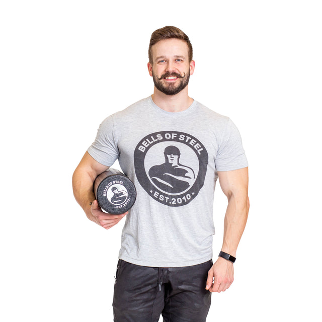 A man in a grey Bells of Steel t-shirt holds a black Bells of Steel Foam Roller, ready for mobility and self-myofascial release, standing against a plain white background.