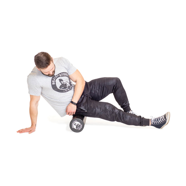 A man uses a Bells of Steel Foam Roller on his right thigh for self-myofascial release, supporting himself with his left hand on the floor. He is dressed in a gray t-shirt, black pants, and black sneakers.