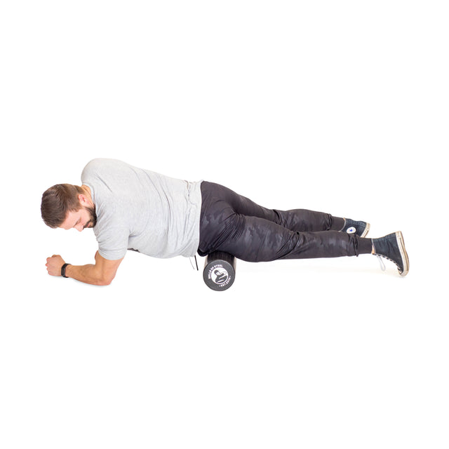 A man uses a Bells of Steel Foam Roller under his right thigh for self-myofascial release, supporting himself on his forearms while lying on his side, dressed in a gray t-shirt and black pants against a white background.