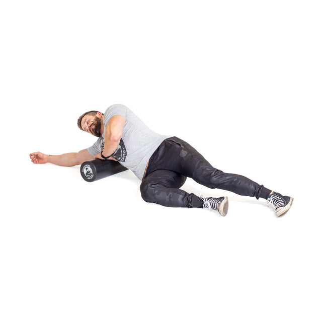 A man wearing a gray t-shirt and black pants smiles as he uses the Bells of Steel Foam Roller for self-myofascial release under his upper arm, stretching on a white background.