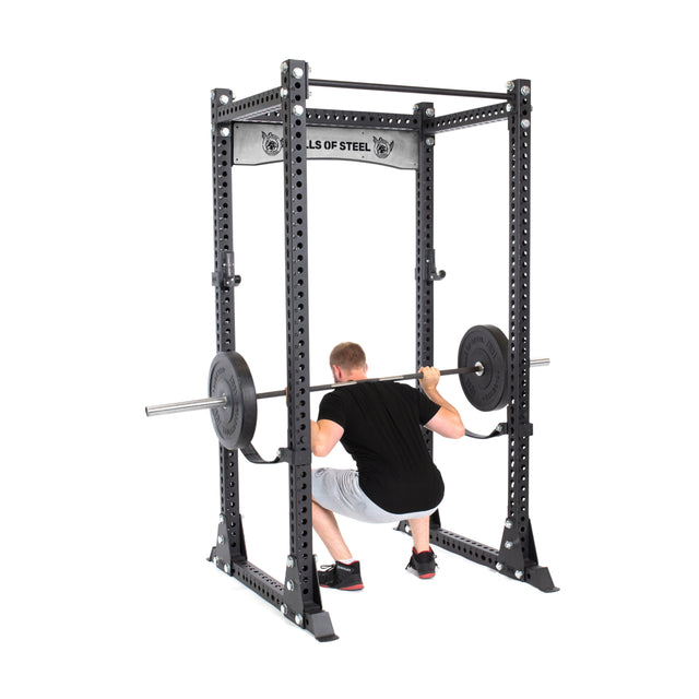 A person squats with a barbell in a black Bells of Steel Manticore Flat Foot Components power rack, wearing a black shirt, gray shorts, and sneakers. The sturdy rack features multiple holes and displays the "Bells of Steel" label at the top for reliable flat foot support during lifts.