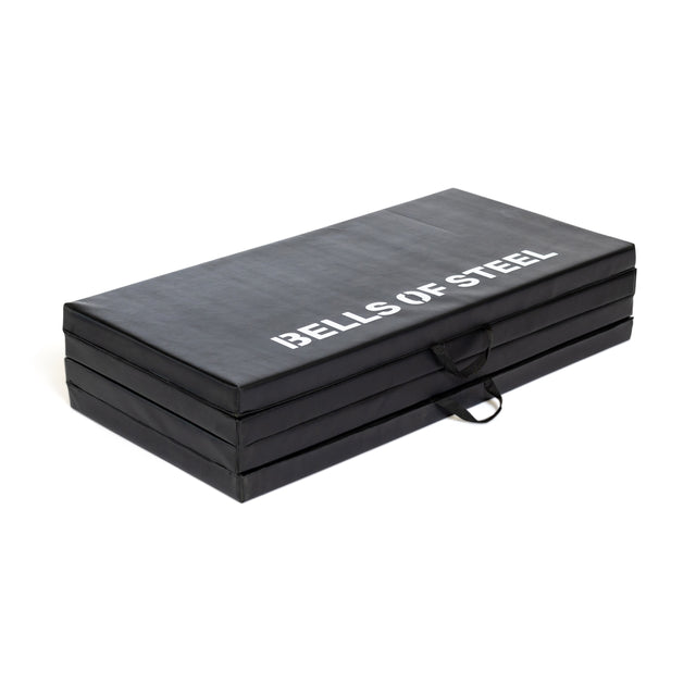 The Bells of Steel Four Fold Mat is a black, foldable plush foam exercise mat with white branding on top. It features four stackable sections and carrying handles for portability.