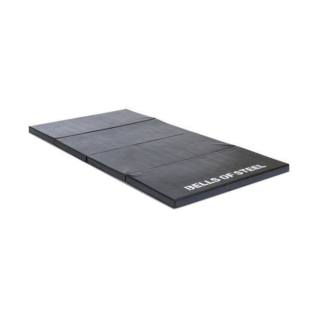 The Four Fold Mat by Bells of Steel is a black, foldable portable gym mat with plush foam and "BELLS OF STEEL" in white text on one end, shown laid flat on a white background.