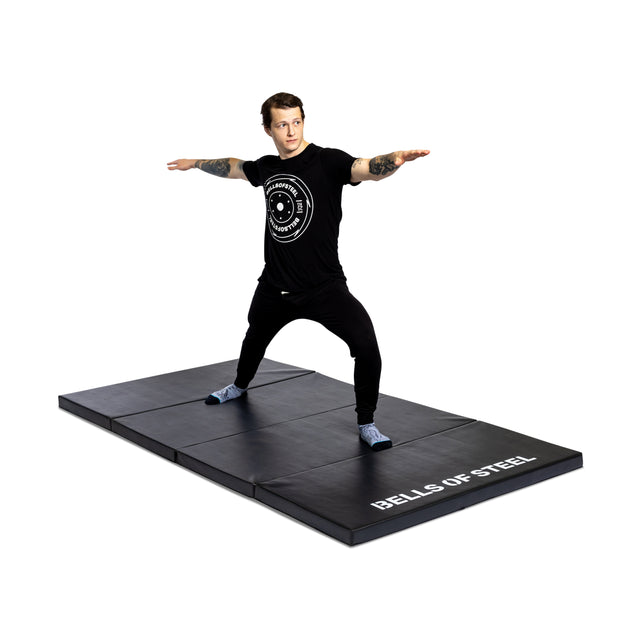 Wearing athletic gear, a person practices a wide-stance pose with arms extended on the large, black, non-slip Bells of Steel Four Fold Mat against a white background.