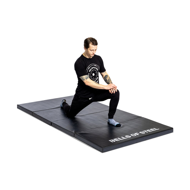 A person in black athletic wear kneels on one knee on a Bells of Steel Four Fold Mat, stretching with hands on their front knee. The background is plain white.