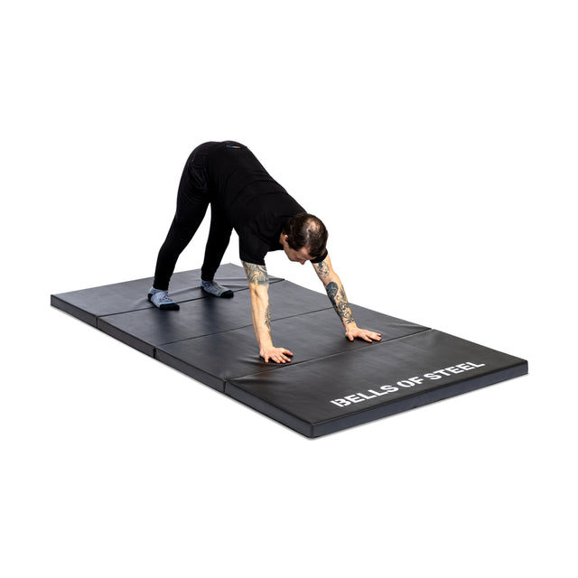 A person in black stretches on a large, non-slip Bells of Steel Four Fold Mat, with both hands and feet touching the surface.