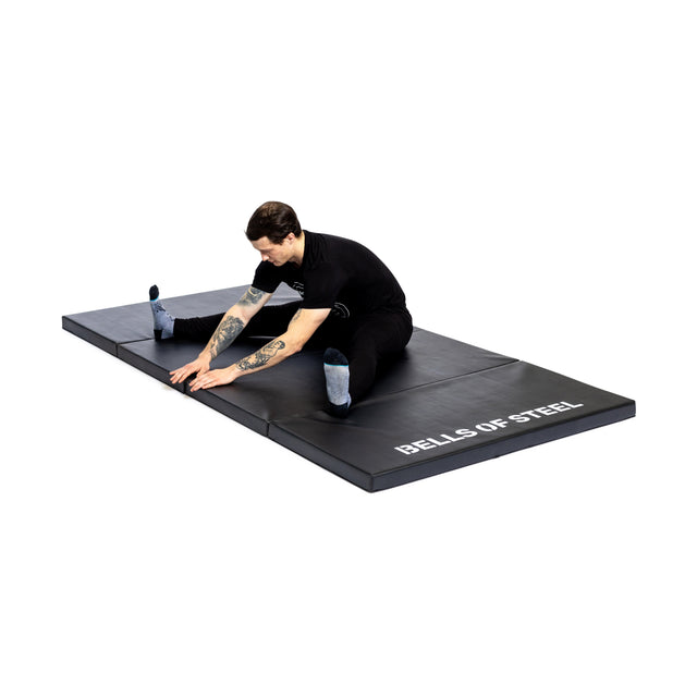 A person in black athletic wear stretches toward one foot while seated on the Bells of Steel Four Fold Mat.