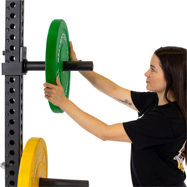 A woman loads a green weight plate onto Bells of Steel Pin Plate Pegs attached to a power rack, with a yellow plate beneath and numbered holes for adjustable accessories, all set against a white background.