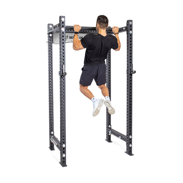 A person uses the Bells of Steel Fat/Skinny Pull Up Bar, performing pull-ups on this sturdy metal rack with multiple adjustment holes and an integrated bar. They wear a black shirt, black shorts, and white sneakers.