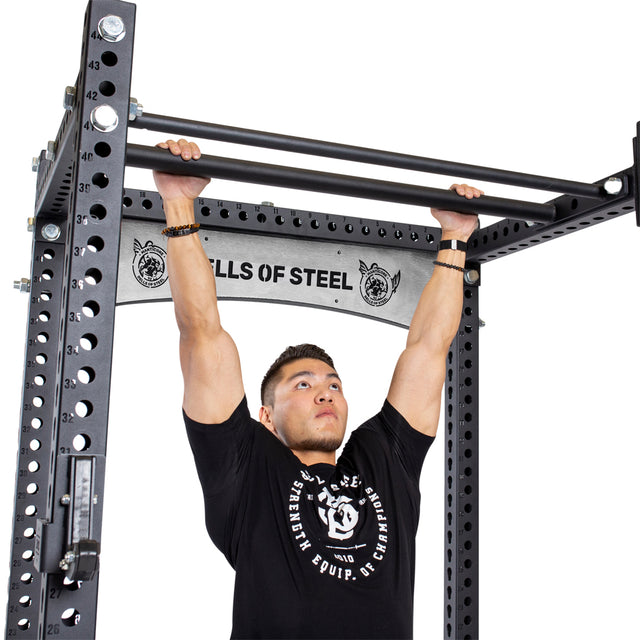 A man in a black t-shirt does a pull-up on the Bells of Steel Fat/Skinny Pull Up Bar, which is attached to a power rack. The "Bells of Steel" brand and a cable pulley are visible in the background.