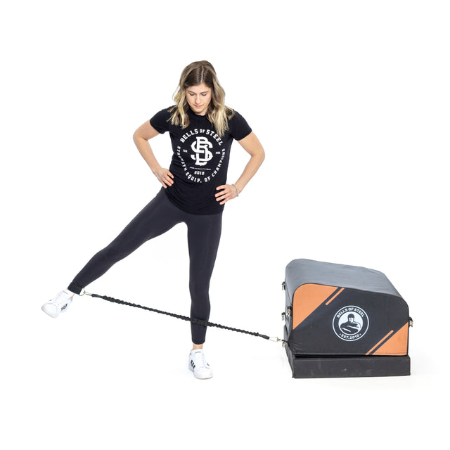A woman uses the Bells of Steel Soft Glute Bench and a resistance band with her left ankle, balancing on her right leg in black attire—showcasing an ideal home workout setup.