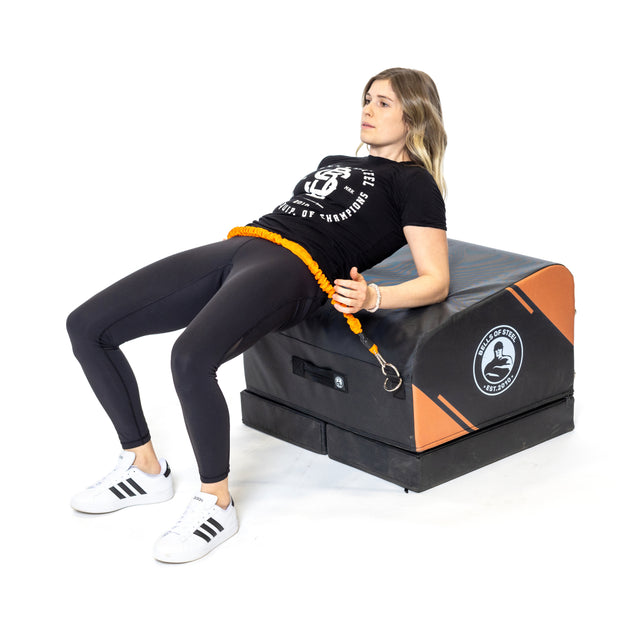A woman does hip thrusts using the Bells of Steel Soft Glute Bench and a resistance band. Dressed in black leggings, a black t-shirt, and white sneakers, she enjoys an ideal home glute workout.