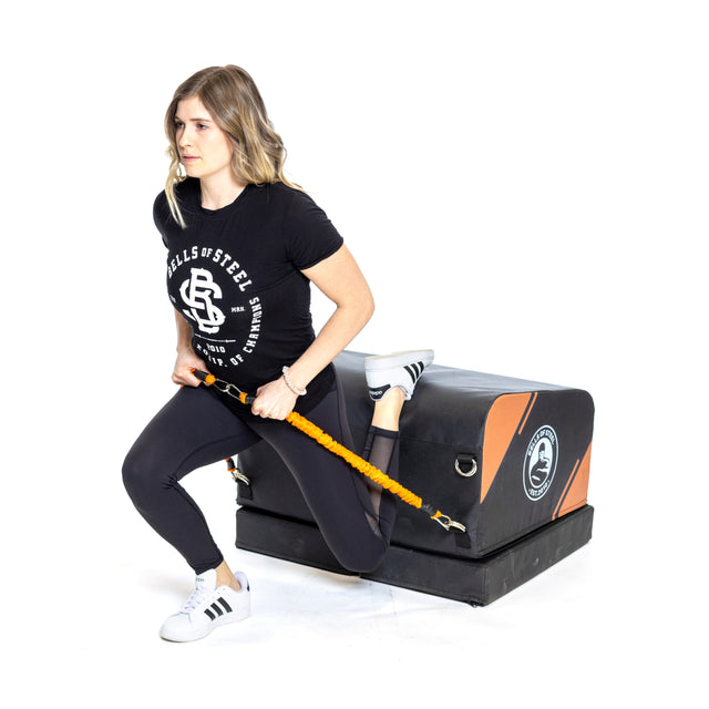 A woman in athletic wear performs a split squat with one leg on the Bells of Steel Soft Glute Bench, holding resistance bands, against a white background.