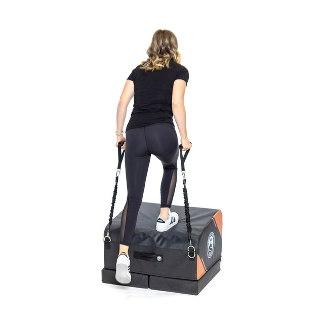 A woman in black athletic wear steps onto the Bells of Steel Soft Glute Bench, holding resistance bands attached to each side, against a white background.