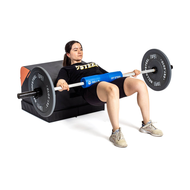 A woman uses the Bells of Steel Soft Glute Bench for a hip thrust exercise, resting her upper back on the bench and lifting a barbell over her hips to power an effective glute workout at home.