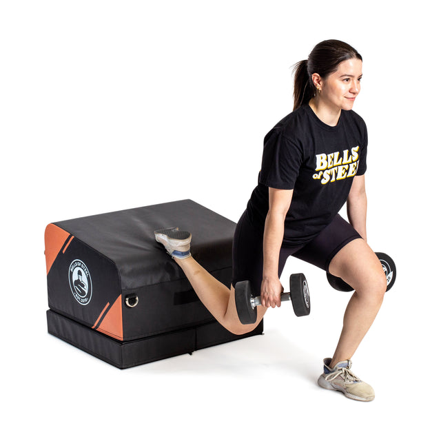A woman does a Bulgarian split squat with dumbbells, her rear foot on the Bells of Steel Soft Glute Bench. She wears a Bells of Steel t-shirt, black shorts, and athletic shoes—ideal for glute workouts at home.