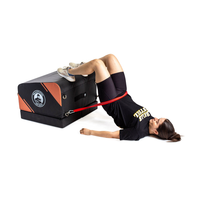 Wearing athletic gear, a woman uses the Bells of Steel Soft Glute Bench and a resistance band for hip thrusts, making it ideal for an effective home glute workout.