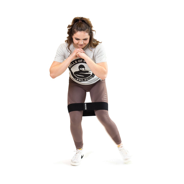 A woman with curly hair exercises laterally with a Bells of Steel Glute Loop resistance band around her thighs, dressed in a gray t-shirt and mauve leggings, smiling as she enjoys her booty workout.