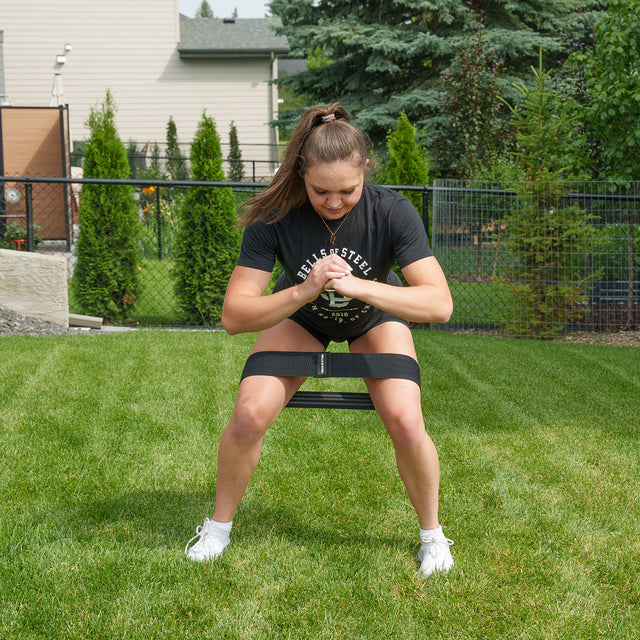 A person outdoors squats on grass with a Bells of Steel Glute Loop above their knees, wearing a black shirt, shorts, and white sneakers. Trees and a fence are in the background.