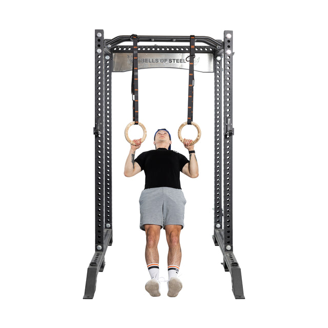 Using Bells of Steel Gymnastics Rings, a person showcases bodyweight training, suspended from a metal frame. They're dressed in a black shirt, gray shorts, blue cap, and sports shoes with socks. This minimal setup highlights the essence of portable fitness.