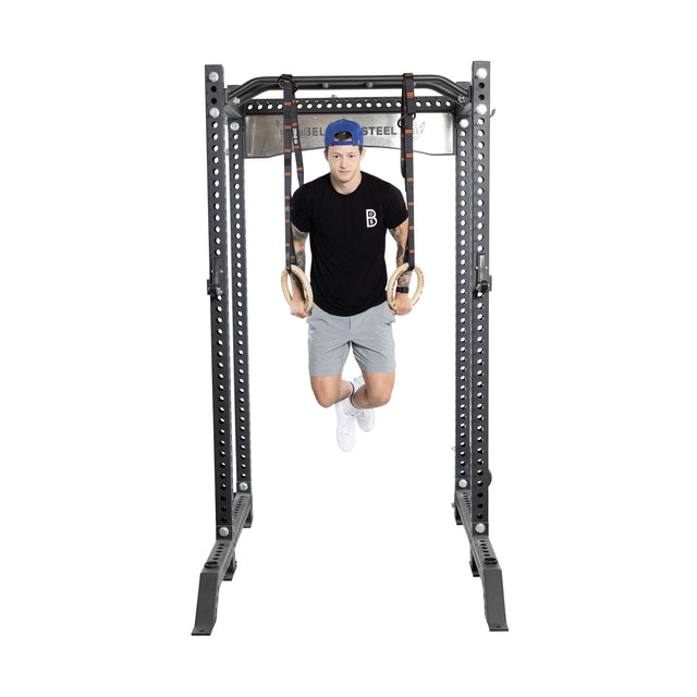 Wearing a blue cap, black T-shirt, and gray shorts, a person performs a dip using Bells of Steel Gymnastics Rings attached to a metal rack against a plain white background.