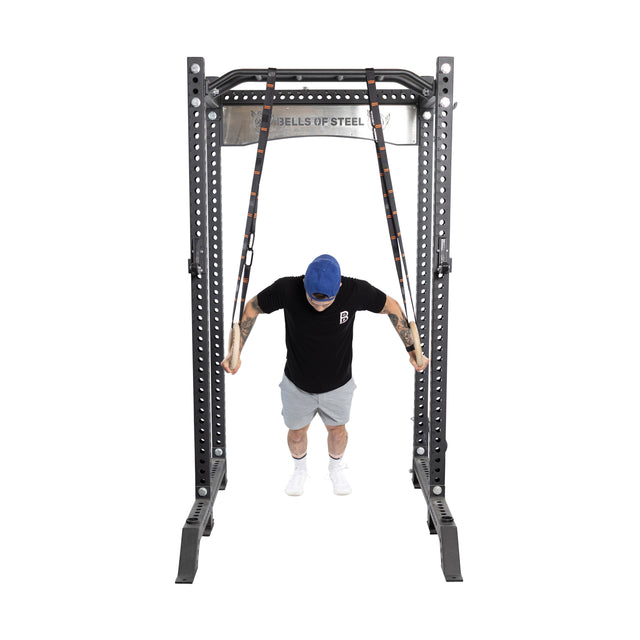 A person in Bells of Steel's black t-shirt and gray shorts, with a blue cap, demonstrates portable fitness using Gymnastics Rings attached to a steel workout rack against a plain white background.