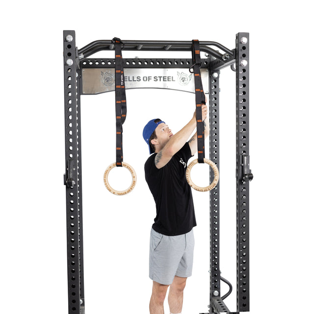 A person in a black shirt and gray shorts adjusts Bells of Steel Gymnastics Rings on a sturdy pull-up bar, perfect for bodyweight training and offering versatile options for portable fitness enthusiasts.