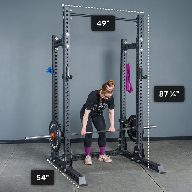 A person lifts a barbell inside the Bells of Steel Manticore Half Rack - Prebuilt (3" x 3", 1" Holes), a sturdy black power rack with robust 3x3 uprights. Clearly labeled dimensions show the rack is 49" wide, 54" deep, and 87¼" tall. A purple band and blue grips hang from the rack, making it a versatile centerpiece for any home gym.