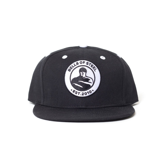 The Bells of Steel Snap Back Hat is a black flat-brim snapback featuring a round white front patch with the "BELLS OF STEEL EST. 2010" logo and strongman graphic, made from durable acrylic wool for comfort and style.
