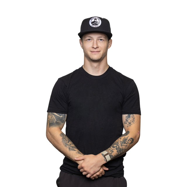 A young man in a black t-shirt wears the Bells of Steel Snap Back Hat, featuring a circular white logo, as he stands against a white background with arms loosely crossed, tattoos visible on both forearms, and a slight smile at the camera.