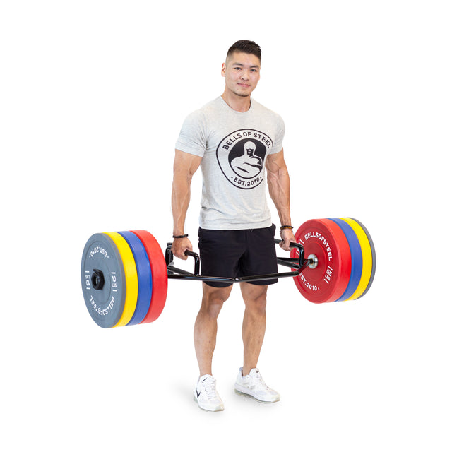 Wearing a gray Bells of Steel shirt and black shorts, a person lifts the Bells of Steel Open Trap Bar / Hex Bar loaded with colorful plates. The bar’s rotating sleeves provide smooth movement, set against a plain white background.