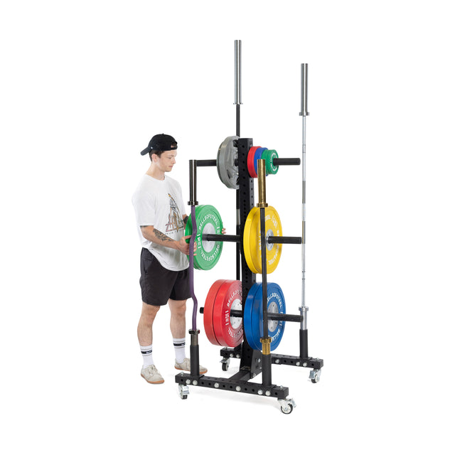A person in a white T-shirt, black shorts, and cap adjusts weight plates on a Bells of Steel Hydra Bumper Plate Tree Builder. The rack is perfect for any home gym and holds various colored plates with two barbells against a plain white background.