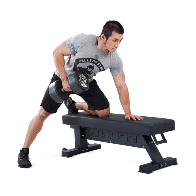Wearing athletic gear, a man kneels with his left knee on the Bells of Steel Hero Heavy-Duty Weight Bench, supports himself with his left hand, and performs a single-arm dumbbell row with his right arm.