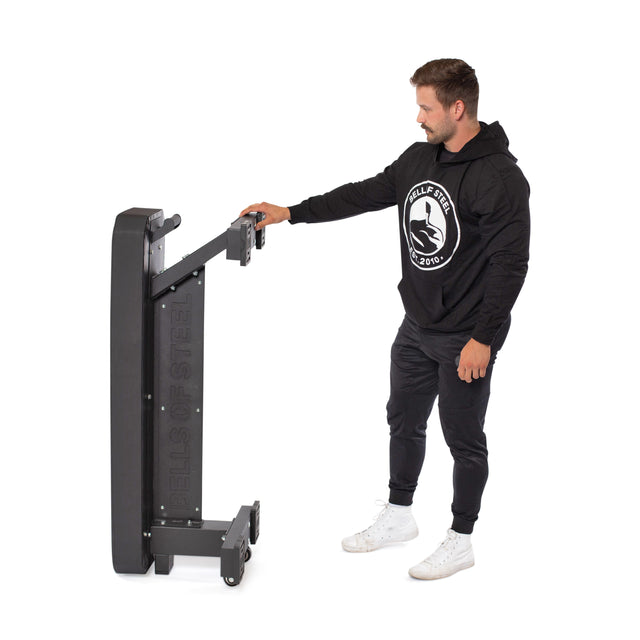 A man in black athletic clothes and white sneakers stands beside a folded Hero Heavy-Duty Weight Bench by Bells of Steel (1,000lb max capacity) against a white background.