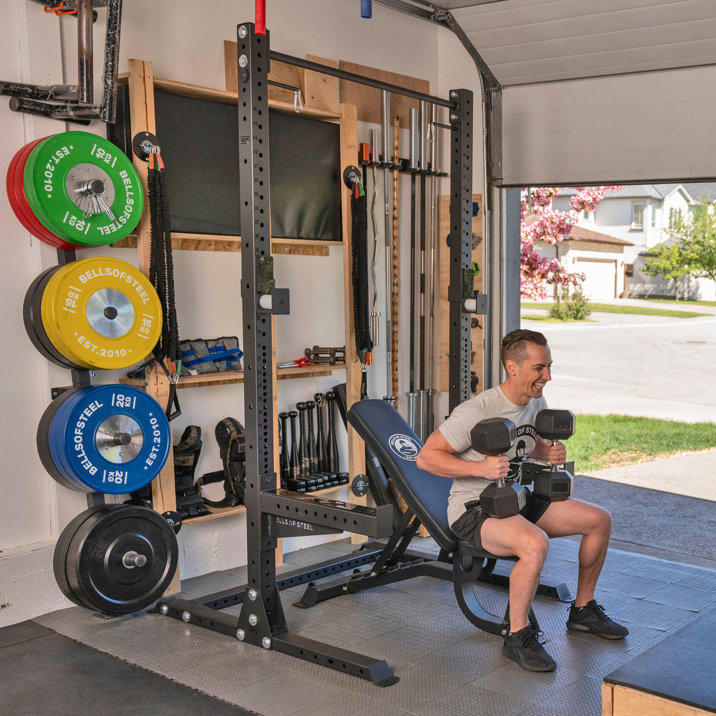 A man uses the Bells of Steel USA Home Gym Builder adjustable bench in his garage gym, surrounded by organized weights and barbells. Sunlight enters through the open garage door, with a suburban street visible outside.