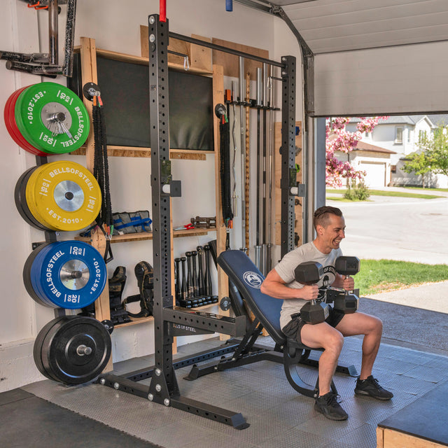 A man uses the Bells of Steel USA Home Gym Builder adjustable bench in his garage gym, surrounded by organized weights and barbells. Sunlight enters through the open garage door, with a suburban street visible outside.